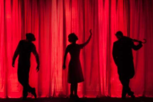 Three performers silhouetted against a lit crimson theater curtain
