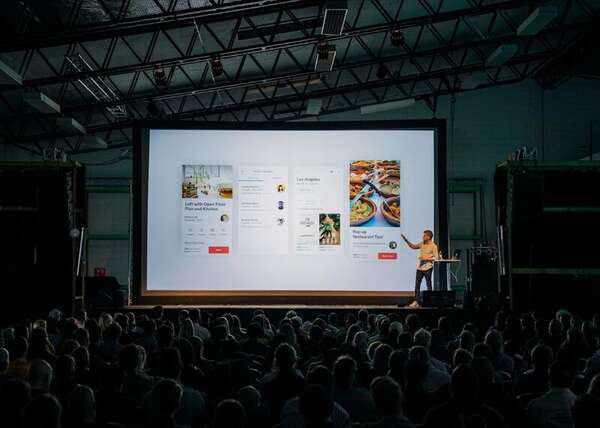 Keynote speaker gesturing at a large booking-interface screen before a silhouetted audience